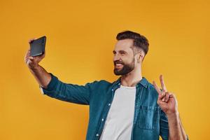 Charming young man in casual clothing taking selfie smart phone and smiling while standing against yellow background photo