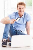 compras en casa. un joven apuesto que trabaja en una laptop y muestra una tarjeta de crédito mientras está sentado en el suelo de su apartamento foto