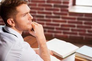 He needs some fresh ideas. Top view of handsome young man in shirt and tie holding hand on chin and looking away while sitting at his working place photo