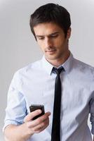 Man typing business message. Handsome young man in shirt and tie holding mobile phone and looking at it while standing against grey background photo