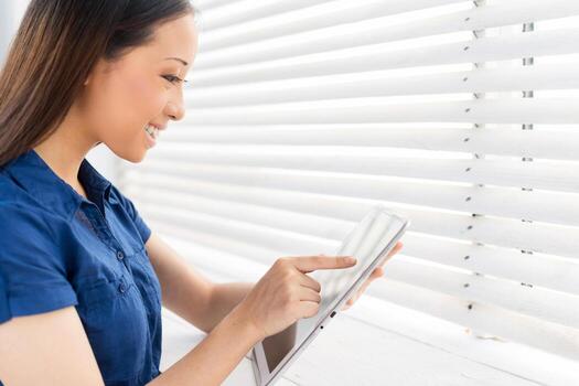 Examining her brand new tablet. Side view of cheerful young Asian woman working on digital tablet and smiling while standing in front of the window photo
