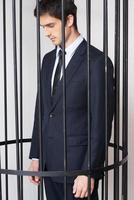 Business criminal. Depressed young man in formalwear standing behind a prison cell and looking at away photo