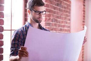 Architect examining blueprint. Confident young man in glasses examining blueprint while standing near the window photo