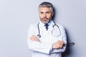 Confident doctor. Mature grey hair doctor looking at camera and keeping arms crossed while standing against grey background photo
