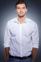 Confident young man. Handsome young man in white shirt looking at camera and holding hands in pockets while standing against grey background photo