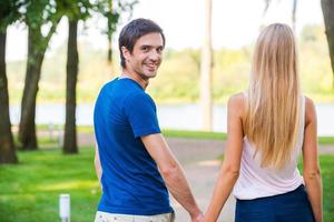 feliz de estar con ella. vista trasera de un apuesto joven mirando por encima del hombro y sonriendo mientras camina por la carretera del parque con su novia foto