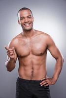 Confident in his perfect body. Young shirtless African man pointing away and smiling while standing against grey background photo