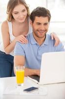 navegando juntos por la red. un joven apuesto sentado en la mesa y usando una laptop mientras su novia está detrás de él