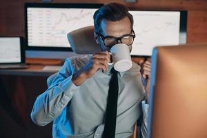 Confident young man drinking coffee and working on computer while staying late in the office photo