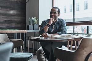 Young and smart. Good looking young man keeping hand on chin and looking away while sitting at his working place in the cafe photo