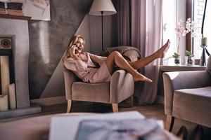 Happy to talk with friend.  Attractive young woman in elegant dress talking on the smart phone and smiling while sitting in the armchair photo