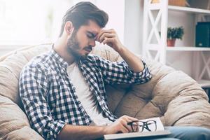 Feeling tired. Frustrated young man holding book and massaging his nose while sitting in big chair at home and keeping eyes closed photo