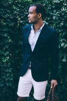 Man in style. Handsome young African man in smart casual wear carrying bag and looking away while standing against green plant background outdoors photo