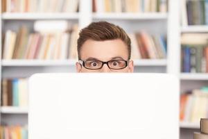 Shocking discovery. Shocked young nerd man in glasses looking out of computer while sitting at the library photo