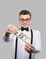 sus primeras ganancias. un joven alegre con corbata de moño y tirantes sosteniendo un billete de cien dólares y sonriendo mientras se enfrenta a un fondo gris foto