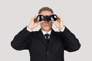 Looking for the new opportunities. Confident mature man in formalwear looking through binoculars while standing against grey background photo