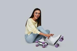 Sincere fun. Beautiful young Asian woman tying roller skates and smiling while sitting against grey background photo