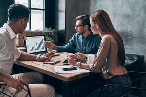 grupo de jóvenes en ropa casual inteligente analizando datos usando una computadora portátil mientras trabajan en la oficina foto