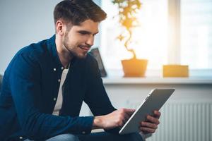 Technologies bring joy. Side view of handsome young man using his digital tablet with smile while sitting on the couch at home photo
