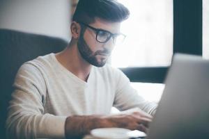 hombre de negocios confiado en el trabajo. un joven confiado con anteojos trabajando en una laptop mientras está sentado en la oficina o en la cafetería foto