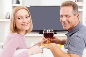felices de estar juntos. vista trasera de una pareja madura sonriente sentada frente a la televisión y sosteniendo vasos con vino tinto foto