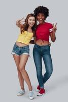 Full length of two attractive young women looking at camera and smiling while standing against grey background photo