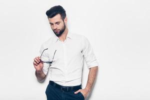 Confident in his style. Handsome young man in white shirt holding sunglasses and looking at them while standing against white background photo