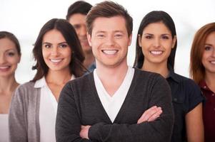 He is a real team leader. Confident young man keeping arms crossed and smiling while group of people standing on background photo