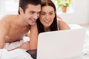 Surfing web together. Cheerful young loving couple lying in bed and using computer together photo