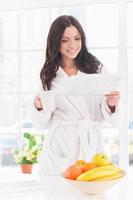 Reading morning news. Beautiful young smiling woman in bathrobe reading newspaper with cup of coffee photo