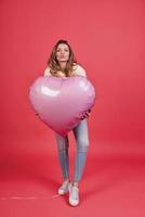 Crazy party girl.   Full length of playful young woman holding heart shaped balloon and puckering while standing against pink background photo