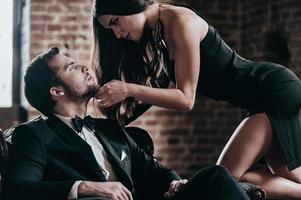 Unleashed desire. Beautiful young woman in cocktail dress leaning to her boyfriend sitting in chair while looking at each other in loft interior photo