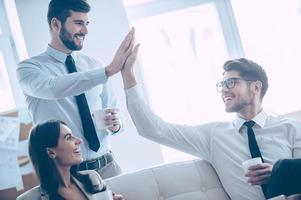 Give me high-five  Two handsome man giving high-five and holding coffee cups with smile while sitting on the couch at office with their beautiful coworker photo