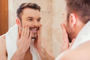 Taking good care of his face. Handsome young man touching his face and smiling while standing in front of the mirror photo