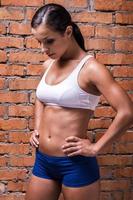 She is in perfect form. Beautiful young woman in sports clothing holding hands on hip and looking down while standing against brick wall photo