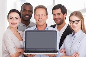 Happy business team. Group of cheerful business people in casual wear standing close to each other while mature man showing laptop monitor and smiling photo