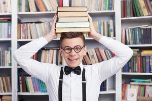 le encanta estudiar. un joven alegre con camisa y corbata de moño sentado en la mesa de la biblioteca y sosteniendo una pila de libros en la cabeza foto