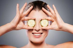 Natural spa treatment. Cheerful mature woman holding pieces of cucumber in front of her eyes while standing isolated on white background photo