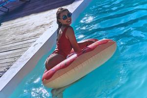 atractiva mujer joven en traje de baño sonriendo mientras está de pie en el anillo inflable en la piscina foto