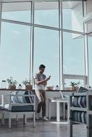 Beautiful day for new achievements. Handsome young man typing something using a smart phone while leaning on the window sill in restaurant photo