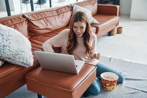 Attractive young woman using laptop and eating popcorn while spending time at home photo