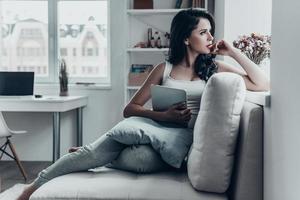 Lazy day. Thoughtful young woman holding digital tablet and sitting on couch while looking outside through the window photo