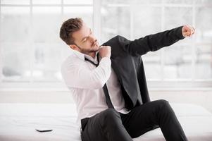 Getting ready for new working day. Young beard man dressing up his jacket while sitting on the bed photo