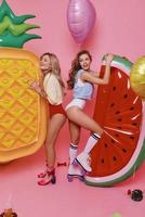 Pure happiness. Full length of two attractive young women in swimwear playing with fruit decorations and smiling while standing against pink background photo