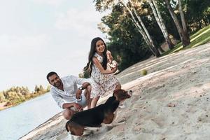 Enjoying great sunny day. Happy young family playing with dog and smiling while spending time on the lake shore photo