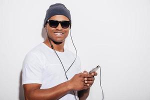 disfrutando de su música favorita. apuesto joven africano con auriculares sosteniendo su teléfono inteligente y sonriendo mientras está de pie contra el fondo blanco foto