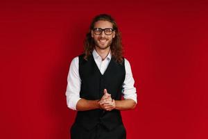 Handsome young man in elegant clothing looking at camera and smiling while standing against red background photo