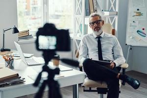 Confident mature man in elegant shirt and tie telling something while making social media video photo