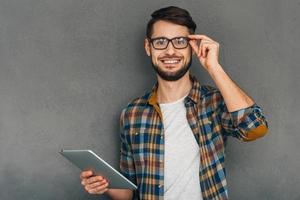 Can I help you Cheerful young man in glasses holding his digital tablet and looking at camera with smile while standing against grey background photo