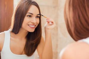 Everything should be perfect. Attractive young woman doing make-up while looking at the mirror and smiling photo